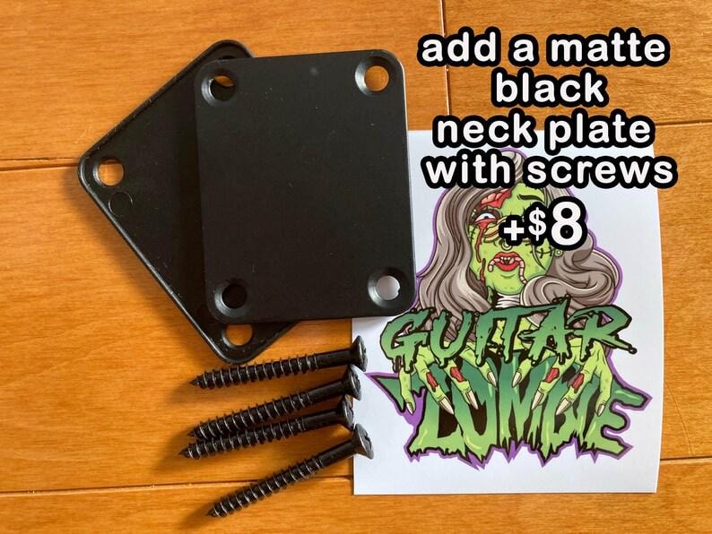 May include: Two black metal neck plates with screws for a guitar. The plates are on a wooden surface. A sticker with a zombie hand and the text "Guitar Zombie" is in the background. The text "add a matte black neck plate with screws +&pound;8" is also visible.