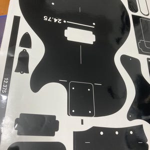 Puede incluir: Una hoja de calcoman&iacute;as de vinilo negro con el contorno de un cuerpo de guitarra, una forma de clavijero y varias plantillas de componentes. El dise&ntilde;o incluye marcas de texto en blanco que dicen 24.75 y 12.375, indicando medidas en pulgadas.