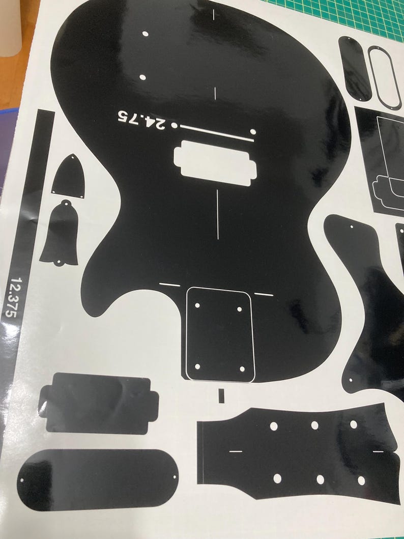 Puede incluir: Una hoja de calcoman&iacute;as de vinilo negro con el contorno de un cuerpo de guitarra, una forma de clavijero y varias plantillas de componentes. El dise&ntilde;o incluye marcas de texto en blanco que dicen 24.75 y 12.375, indicando medidas en pulgadas.