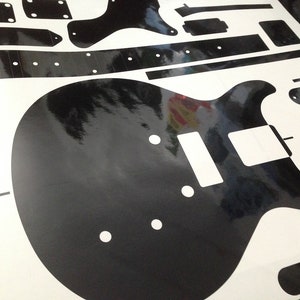 Puede incluir: Un conjunto de calcoman&iacute;as de vinilo negro que presenta la silueta del cuerpo de una guitarra el&eacute;ctrica, una tira larga para el diapas&oacute;n con marcadores circulares y varios componentes rectangulares y geom&eacute;tricos sobre una hoja blanca.