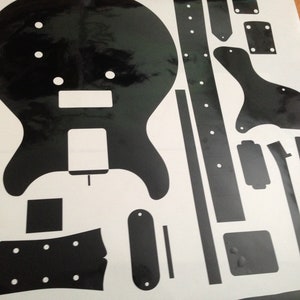 Puede incluir: Una hoja de pegatinas de vinilo negro con varios componentes de guitarra, incluyendo una silueta de cuerpo s&oacute;lido, una forma de clavijero, una tira de diapas&oacute;n, un golpeador y varias formas rectangulares y ovaladas peque&ntilde;as sobre fondo blanco.