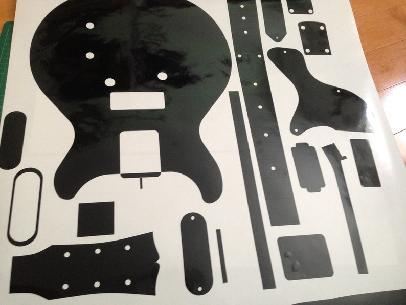 Puede incluir: Una hoja de pegatinas de vinilo negro con varios componentes de guitarra, incluyendo una silueta de cuerpo s&oacute;lido, una forma de clavijero, una tira de diapas&oacute;n, un golpeador y varias formas rectangulares y ovaladas peque&ntilde;as sobre fondo blanco.