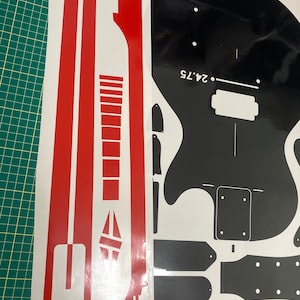 Puede incluir: Un conjunto de calcoman&iacute;as de vinilo adhesivo que incluye una tira roja larga con formas geom&eacute;tricas y una hoja negra con la silueta del cuerpo de una guitarra el&eacute;ctrica, un clavijero y varios componentes con el texto 24.75 impreso.