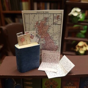 May include: A miniature book with a blue cover and a stack of old letters on top of it. The book is sitting on a wooden shelf with other books. Behind the book is a map of the British Isles titled "Atlas of the World".