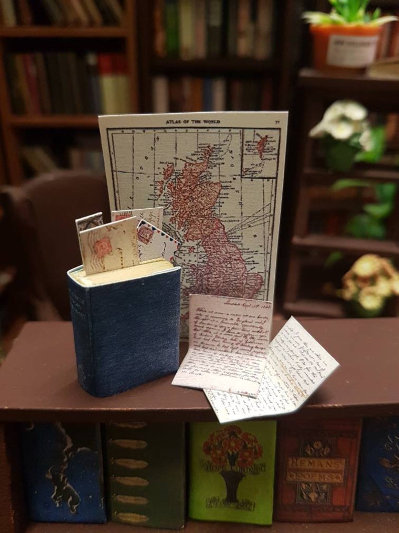 May include: A miniature book with a blue cover and a stack of old letters on top of it. The book is sitting on a wooden shelf with other books. Behind the book is a map of the British Isles titled "Atlas of the World".