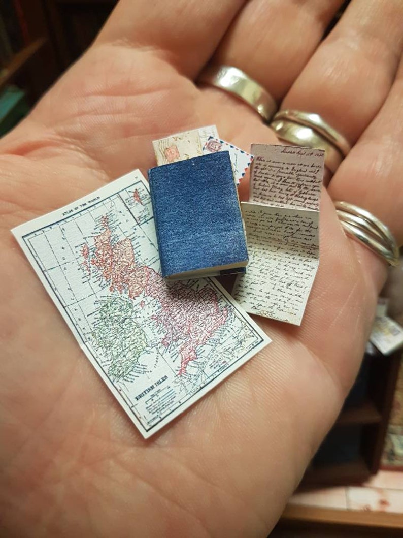 May include: A miniature map of the British Isles, a small blue book, and a handwritten letter are displayed on a hand. The map is titled "British Isles" and has a grid pattern. The letter is dated "London, Aug 17, 1893".