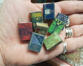 May include: A collection of miniature books with various titles and colorful covers. The books are arranged on a hand, with the titles visible. The titles include "Bob Burton", "The Ingoldsby Legends", "Tales From Dickens", and "The  Mysterious Traveller".