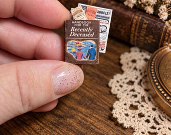 May include: A miniature book titled Handbook for the Recently Deceased with a brown cover featuring a graphic illustration of two figures standing before a landscape. Behind the book are small printed papers including one labeled EXORCIST.