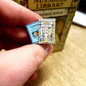 May include: Two tiny books from the Nutshell Library by Maurice Sendak are held in a hand. One is light blue and titled "Pierre". The other is yellow and features a drawing of a crocodile and a child. A larger box with the title "Nutshell Library" is in the background.