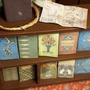 May include: A miniature bookshelf with a variety of books, including a book titled "Hemans' Poems" and a map titled "The Shire". The bookshelf is made of wood and has a brown finish. There is a small, brown book on the top shelf, and a green plant is visible in the background.