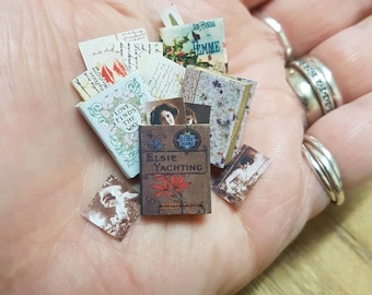 May include: A collection of miniature books and vintage postcards, including titles like "Love Finds the Way" and "Elsie Yachting", displayed in a hand.