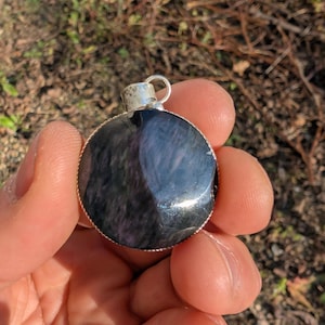 Può includere: Un ciondolo rotondo con un'ansa argentata. Il ciondolo presenta una pietra lucida, grigio scuro, con motivi a vortice. La pietra è incastonata in una lunetta d'argento.