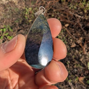 Op de afbeelding: Een druppelvormige hanger met een gepolijste, iriserende steen. De steen toont tinten blauw, groen en paars, gezet in een zilverkleurig metalen frame. De hanger wordt in een hand gehouden tegen een vage buitenachtergrond.