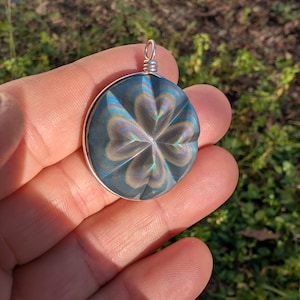 May include: A circular pendant featuring a four-leaf clover design. It has a silver-coloured metal frame and an iridescent centre in shades of blue, green, and silver. The pendant is held in a hand against a blurred green backdrop.