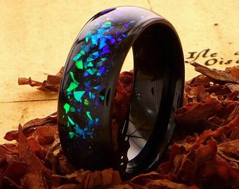 May include: A black ceramic ring with a blue and green iridescent inlay. The ring is sitting on a bed of brown tobacco flakes.