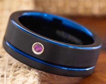 May include: A black ring with a blue interior and a thin blue stripe. The ring features a small, round, pink gemstone. The ring is made of a dark, matte material.