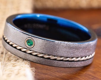 May include: A close-up of a silver ring with a blue interior. The ring features a small green gemstone and a gold-colored rope-like design. The ring is on a wooden surface.