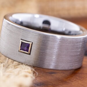 May include: A wide, brushed silver-tone metal ring featuring a single, small, square-cut purple gemstone set in a gold-tone bezel. The band has a flat profile with a textured, matte finish across the outer surface, contrasting with the polished interior.
