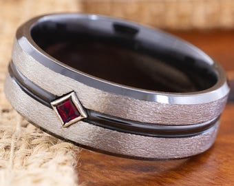 May include: A close-up of a silver-colored ring with a black band and a red gemstone. The ring has a textured finish and a geometric design. The gemstone is a small, square-shaped ruby.