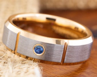 May include: A gold and silver-tone ring with a small blue gemstone. The ring has a brushed silver band with gold accents and a gold interior. The blue gemstone is set in a gold bezel.