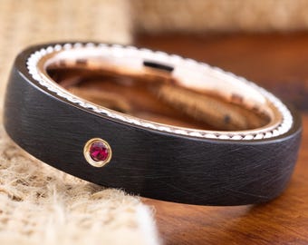 May include: A black ring with a brushed texture, featuring a small, round, red gemstone set in a gold-colored bezel. The ring's interior is a rose gold color, and the edge is detailed with a white, textured pattern. The ring is displayed on a textured surface.