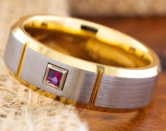 May include: A gold and silver-tone ring with a square, red gemstone. The ring has a brushed silver band with gold accents and a gold interior. The gemstone is set in a square gold frame. The ring is on a textured surface.