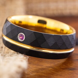 Puede incluir: Un anillo negro con una superficie facetada y un interior dorado. El anillo tiene una fina banda dorada y una pequeña gema rosa redonda. El anillo se muestra sobre una superficie de madera.