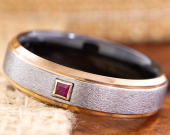 May include: A close-up of a ring with a textured silver band, a gold-colored inner and outer edge, and a black interior. A small, square, red gemstone is set into the silver band. The ring is resting on a textured surface.