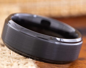 May include: A black ring with a brushed metal finish. The ring has a flat top and a rounded inner edge. The ring is displayed on a wooden surface with a woven fabric background.
