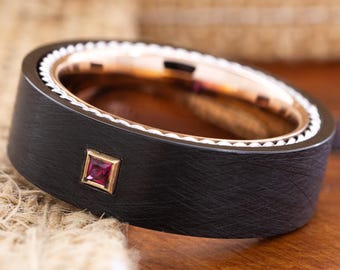 May include: A black ring with a brushed texture, featuring a gold interior and a small, square ruby gemstone. The ring has a white, decorative border and is set against a wooden surface.