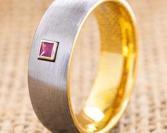 May include: A silver-colored ring with a gold interior and a small, square, ruby-colored gemstone. The ring has a brushed metal finish and a polished gold inner band. The gemstone is set in a square bezel.