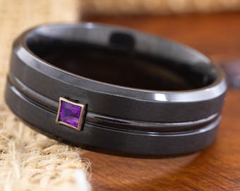 May include: A black ring with a square purple gemstone set in a gold-colored bezel. The ring has two parallel grooves and a matte finish. The ring is displayed on a textured surface.