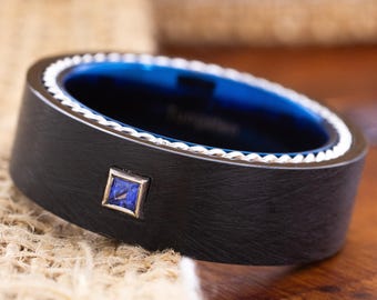 May include: A black ring with a blue interior and a square blue gemstone. The ring has a white decorative border and is set against a neutral background. The ring is a close-up shot.