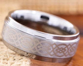 May include: A silver-toned ring with a Celtic knot design etched around the band. The ring has a polished, shiny top and a matte finish on the band with the intricate pattern. The ring is set against a neutral background.