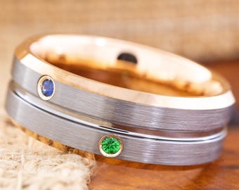 May include: A two-tone ring with a brushed silver band and a gold-colored interior. The ring features a blue gemstone and a green gemstone. The ring is displayed on a wooden surface.