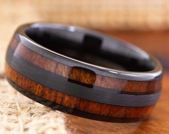 May include: A black ring with a wood inlay and a black center band. The wood has a reddish-brown color and a glossy finish. The ring is on a wooden surface.