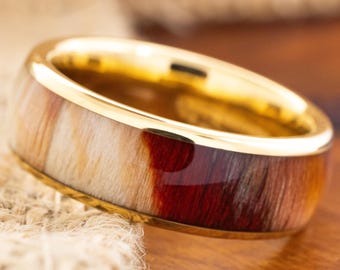 May include: A gold-colored ring with a wide band. The band features a natural wood grain pattern in shades of cream, brown, and deep red. The ring is set against a neutral background, highlighting its design and craftsmanship.
