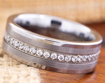 May include: A silver-colored ring with a brushed metal finish. The ring features a row of small, clear gemstones set between two polished metal bands. The ring is displayed on a wooden surface.