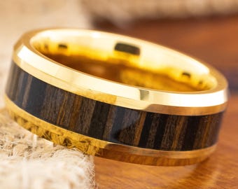May include: A gold-colored ring with a dark wood inlay. The ring has a polished finish and a band of dark brown wood with vertical grain. The ring is displayed on a wooden surface with a burlap texture in the background.