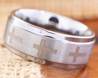 May include: A silver-colored ring with three cross designs etched around the band. The ring has a brushed metal finish and a polished edge. The ring is displayed on a wooden surface.