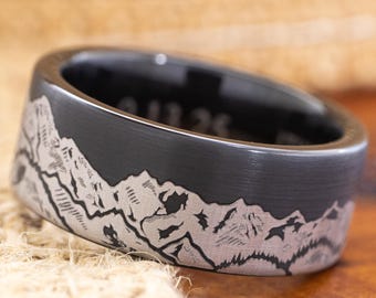 May include: A black ring with a mountain range design etched around the band. The ring has a flat profile and a matte finish. The mountains are detailed with lines and shading, creating a textured appearance. The ring is likely made of metal.