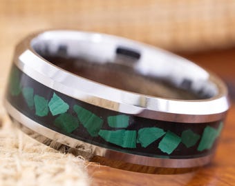 May include: A stainless steel ring with a black inlay filled with irregularly shaped green malachite stones. The ring has a polished, reflective surface and a wide band design. The ring is set against a neutral background.