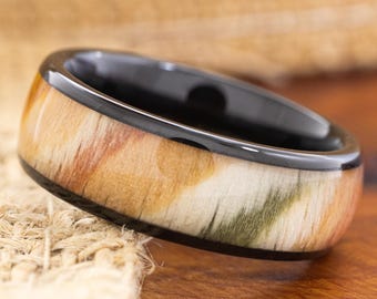 May include: A black ring with a wood grain pattern in shades of brown, tan, and green. The ring has a glossy black band and a natural, organic appearance. The ring is a unique piece of jewelry.