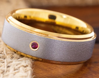 May include: A silver and gold wedding band with a small red gemstone set in the center.