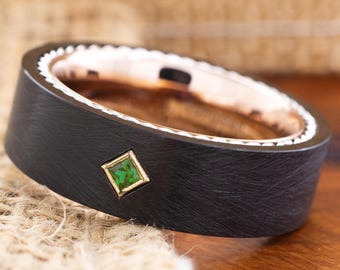 May include: A black ring with a brushed metal finish. The ring features a square, emerald-green gemstone set in a gold bezel. The inner band is rose gold, and the outer edge has a decorative white pattern.