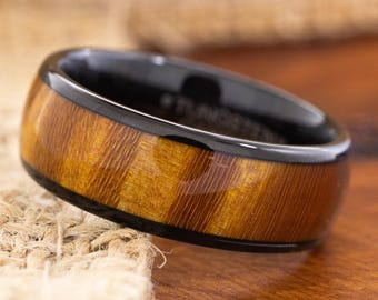 May include: A close-up of a black and wood-tone ring. The ring has a black band with a wood-grain pattern around the center. The ring is made of tungsten. The ring is on a wooden surface.