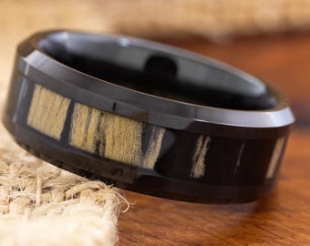 May include: A black ring with a wood inlay. The ring features a black band with a rectangular wood grain pattern in shades of tan and black. The ring is set against a neutral background.