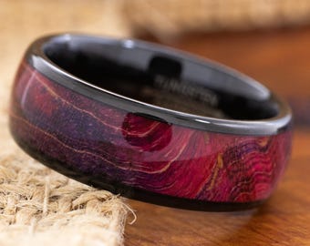 May include: A close-up of a ring with a black band and a vibrant, reddish-purple wood grain inlay. The ring has a polished finish, showcasing the natural patterns and colors of the wood. The ring is a stylish accessory.