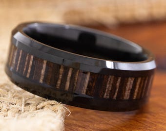May include: A black ring with a wood inlay. The ring has a polished black band with a dark wood grain pattern around the center. The ring is sitting on a wooden surface.