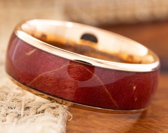 May include: A close-up of a gold-toned ring with a wide band. The ring features a deep red, wood-grain inlay. The gold band has a polished finish. The ring is set against a neutral background.
